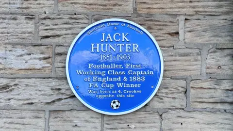 BBC / Mia Fitzpatrick A blue commemorative plaque is mounted on a textured stone wall. The plaque honors Jack Hunter (1851–1903), a footballer and FA Cup winner. It notes that he was the first working-class captain of England and marks his birthplace nearby.
