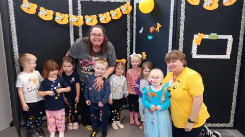 A group of eight children, all under the age of four, stand in a line with two pre-school workers. They all have stickers on their faces and clothes and are standing in front of a banner with Pudsey bear faces on it.