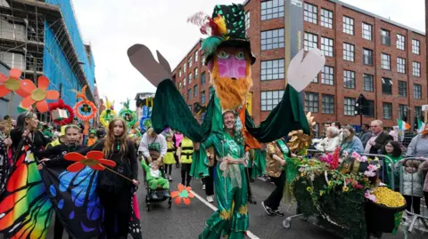 People in colourful costumes walk down a street. In the centre of the street, there is a man in a green silk costume, which includes a cardboard statue of a bearded man attached to his shoulders.