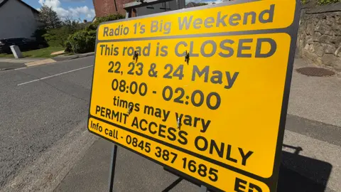 BBC/Jim Scott A yellow road sign warning motorists of the impending road closures. The sign states that the road will be closed on 22, 23 and 24 May, while access will be restricted to those with permits.
