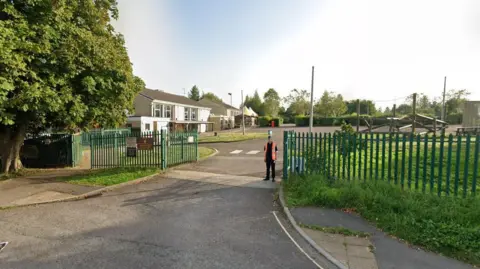 Google Maps Nailsworth Church of England Primary entrance on a sunny day - one storey buildings with a tarmac and grass playground, a large tree to the left of the gates.