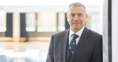 Northampton International Academy Martin Serrão in a navy suit, white shirt and navy tie stands to the left hand side smiling at the camera.