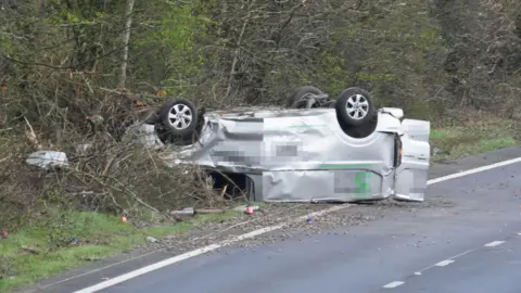 An overturned silver van next to a road. It is sat on its roof in shrubbery by the side of the road.