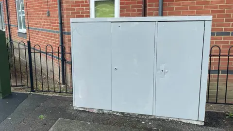 LDRS/UGC A grey utility box box in front of homes which has been painted over with grey paint.