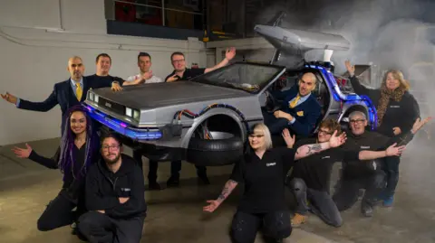 A group of people surround a DeLorean car which appears to be suspended in mid-air partially shrouded in smoke