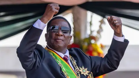 A Getty Images, o presidente do Zimbábue, Emmerson Mnangagwa, com óculos de sol e vestindo um terno e uma faixa nas cores do Zimbábue e uma corrente e uma estrela do escritório segura os punhos.