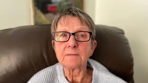 Elderly woman with glasses and a grey jumper on a brown sofa