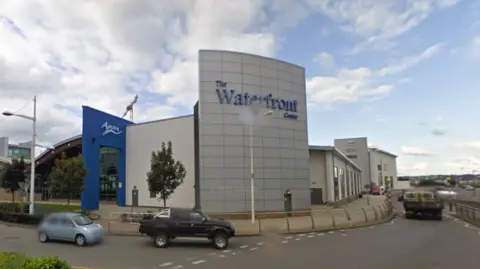 A picture of the front of the leisure centre. It is a large modern building with a main road in front of it. Cars can be seen driving past in the image