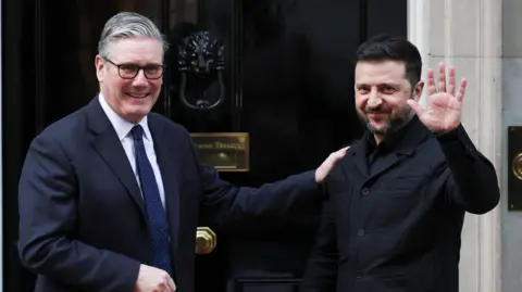 President Volodymyr Zelensky waves at cameras outside the black door of 10 Downing Street as he is welcomed by Prime Minister Sir Keir Starmer, who places his hand on his shoulder.