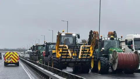 Michael Cooper Several tractors driving in two lanes along a wet road. There are other tractors behind and other cars further behind them.
