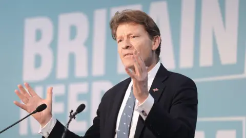 PA Media Richard Tice, wearing a black suit jacket and a union flag lapel badge on his left lapel, is talking to an audience with his hands widened.