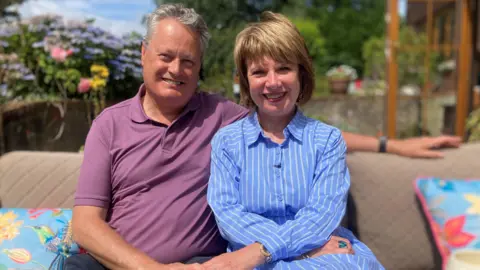 A man and woman are sitting on an outdoor sofa which is covered with soft cushions. The man, who is clean shaven, has short grey hair and is wearing a purple polo neck tshirt. The woman, who is on the right, is wearing a blue and white pinstriped dress. She has medium length blonde hair and is wearing a gold watch and necklace. Both are looking at the camera. Behind them is an out of focus garden showing an open door to the house, a low wall and lots of colourful flowers. 