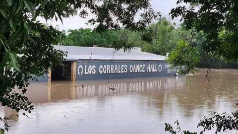 Floodwaters are seen engulfing the Rodeo Los Corrales Dance Hall in Comfort. Trees sit around the hall. 