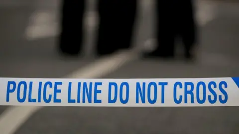 A close up of police tape that reads "police line do not cross".  The blurred legs of two people are in the background.