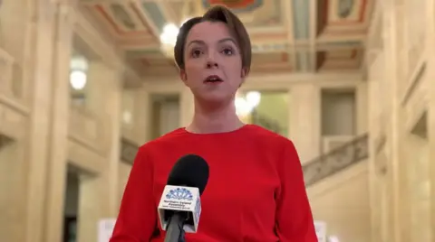 PA A woman with short brown hair is standing a a white marble hall. She is wearing a bright red dress and is talking into a microphone that says 'Northern Ireland Assembly'.