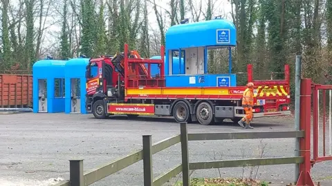 Mike Fuller A blue Humber Bridge toll booth on the back of a flatbed lorry. There are a number of other toll booths standing in front of the lorry.
