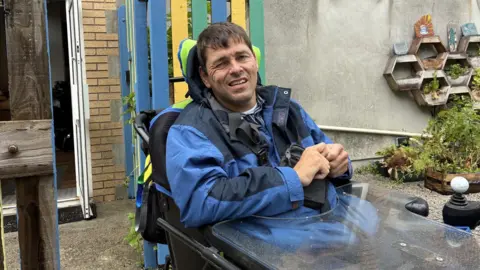 A man in a blue jacket with dark hair smiles at the camera , he uses a motorised wheelchair 