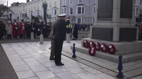 The man is seen standing before wreaths of poppy's with his arm raised to salute. He is stood next to another man taking part in the ceremony as on lookings in the town gather to watch.