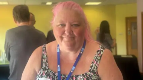 Molly Smith/BBC Tracy Young has pink hair and it wearing a floral top and blue visitor's lanyard. She is smiling and there are two people behind her.