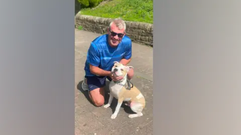 Mick Martindale Facebook A man with grey hair, wearing black sunglasses and a blue shirt and grey shorts, kneels down on the floor, with his hands hugging a Staffordshire bull terrier type dog. It is a sunny day.