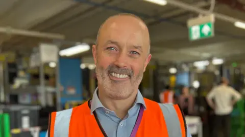 John Boumphrey, Country Manager Amazon UK, standing inside a warehouse. He is in a blue shirt covered by a orange high visibility jacket and is smiling broadly.