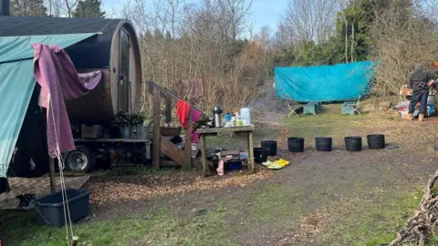 The Stanmer Sauna Garden visible on the left-hand side. A green tarpaulin is propped up to the right of the sauna. Five black buckets can be seen beside a table with drinks. In the distance there are two seats and two benches. 