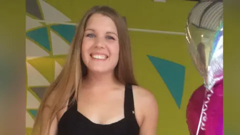 Family supplied Bethany Lilley poses for a picture during her birthday. There is a silver and pink balloon to her left hand side. She is wearing a black strappy top and has long mousey blonde hair. She has blue eyes and is smiling widely. There is a green patterned wall behind her.