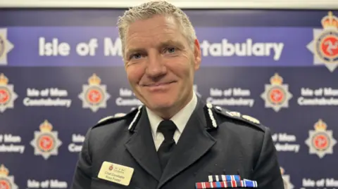 Chief Constable Russ Foster is wearing his black uniform with a white shirt and black tie. He has a slight smile and has short grey hair. He is standing in front of a wall with repeated Isle of Man Constabulary signage across it and the police force crest.