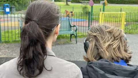 A child wearing headphones next to an adult, with just the back of their heads visible, sitting in a children's playground in a park