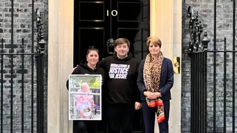 Linsey Smith/BBC Three people are stood in front of Ten Downing Street. They are holding a large framed photograph of a victim of the Legacy Funeral Homes investigation. 
