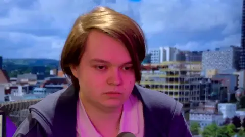 A young man sits in a studio against a purple background