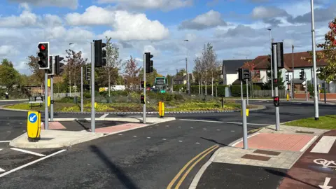 Jozef Hall/BBC A photograph taken from the road approaching a roundabout. A number of sets of traffic lights can be seen, and houses and a green bus can be seen in the distance. 