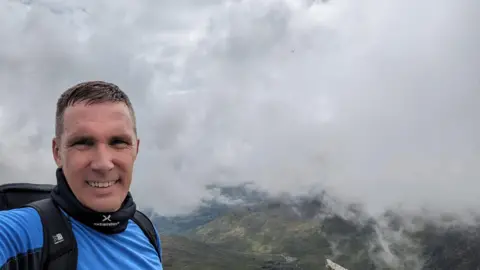 A man in a blue top is standing on Mount Snowdon, the view is visible on the right of the photo