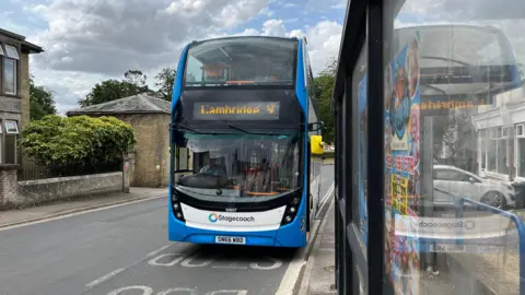 John Devine/BBC A blue Stagecoach double-decker bus at a bus stop in Littleport, there is a bus shelter with glass panels to the right and on the left there are some residential houses.