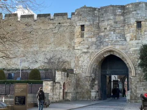 The entrance to Lincoln Castle, a tall beige-coloured stone brick structure. Three people are walking through the archway gate while another man is walking along the pavement towards the front.