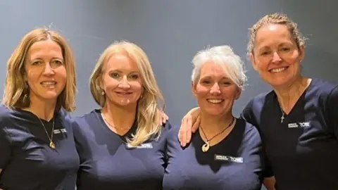 Four women, wearing matching navy blue t-shirts smile as they pose for the camera. They are stood against a grey background, and all four are also wearing necklaces.