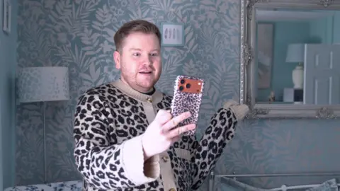 A man in a leopard print cardigan holding a phone in a leopard print case
