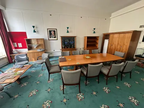 A large office with a big wooden meeting table, chairs and a green carpet emblazoned with lions.