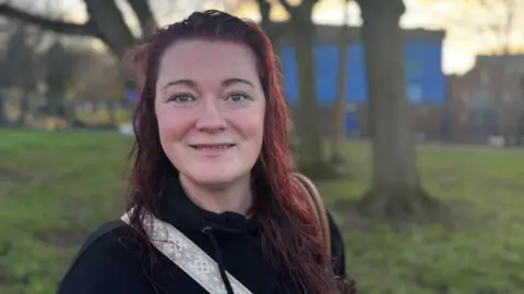 Claire Hamilton/BBC Nicola Ward, who has long dyed red hair and is wearing a black hoodie with the strap of a gym bag over her shoulder, smiles at the camera against the backdrop of a grassy area lined with trees.