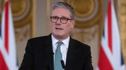 Keir Starmer who has grey hair and dark-rimmed glasses speaks at a podium. He is wearing a dark suit, green tie and white shirt. British flags can be seen in the background.