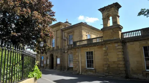 Tony Bartholomew A sandstone building with large rectangular windows and an archway at its centre, with information boards either side. It's surrounded by a blue sky and shrubbery hanging over a black railing.