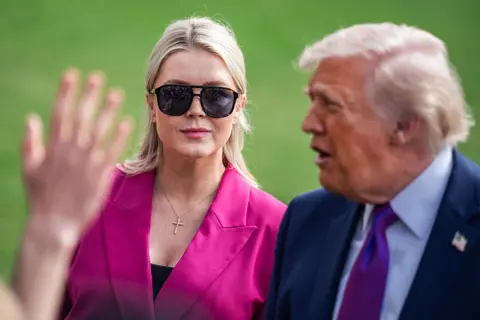 Karoline Leavitt wears sunglasses and a pink blazer as she stands behind Donald Trump during a press gaggle.