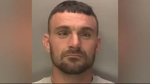 WMP Kasaj has close cropped brown hair and a beard and is wearing a grey T'shirt in this police mugshot.