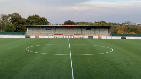 harlow Town football pitch shows the central bit of the grassed area with a circle outlined in white being dissected by a white line. A covered seating stand is in the background with a low fence before it with advertising boards on. 