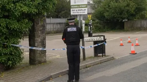 A police officer is seen standing next to a car park with his back to the camera. There are bollards around the area and police tape. 