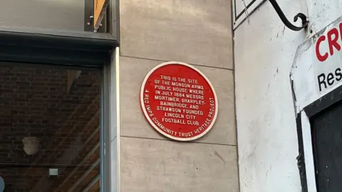 A close up of a red plaque hanging on the wall that says 'THIS IS THE SITE OF THE MANSON ARMS PUBLIC HOUSE WHERE IN JULY 1884 MESSERS MORTIMER SHARPLEY-BAINBRIDGE, AND STRAWSON FOUNDED LINCOLN CITY CLUB.' and around the edge of the plaque it says 'A RED IMPS COMMUNITY TRUST HERITAGE PROJECT'.