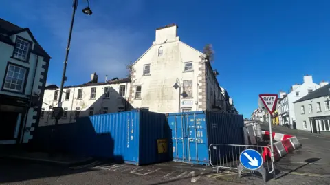 A view of the side of the Antrim Arms Hotel, a white building in a town. Blue storage containers are blocking it off as are bollards and a fence in front of it.