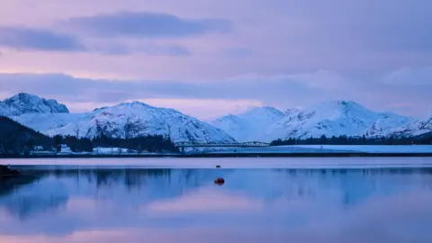 Andrew Briggs A tranquil loch reflects snow-covered peaks under a soft purple and blue winter sky, with a bridge spanning the water in the distance.