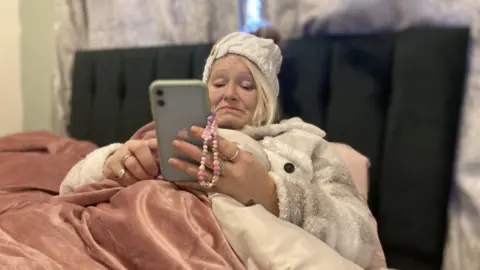 A woman in her 50s is sat in a bed with a pink velvet bedspread and looks at her mobile phone, whch has a grey case. She's wearing a cream-coloured knitted bobble hat and a cream and camel coloured coat. 