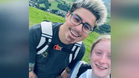 A selfie of Nathaniel and Jade on a walk in the countryside. Nathaniel has bleached hair and wears glasses, while Jade has fair hair tied back in a ponytail.